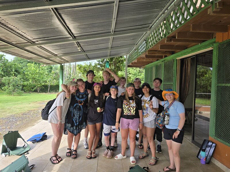 The image shows a group of teenagers posing for a photo under a covered patio. They are of diverse ethnicities and are dressed in casual clothing. Some are holding water bottles or bags. The background reveals greenery and a building with a green door. The overall atmosphere seems relaxed and friendly.